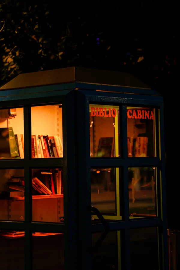A small phone-booth-style free library glows warm orange against the night, books lining its illuminated shelves behind glass.