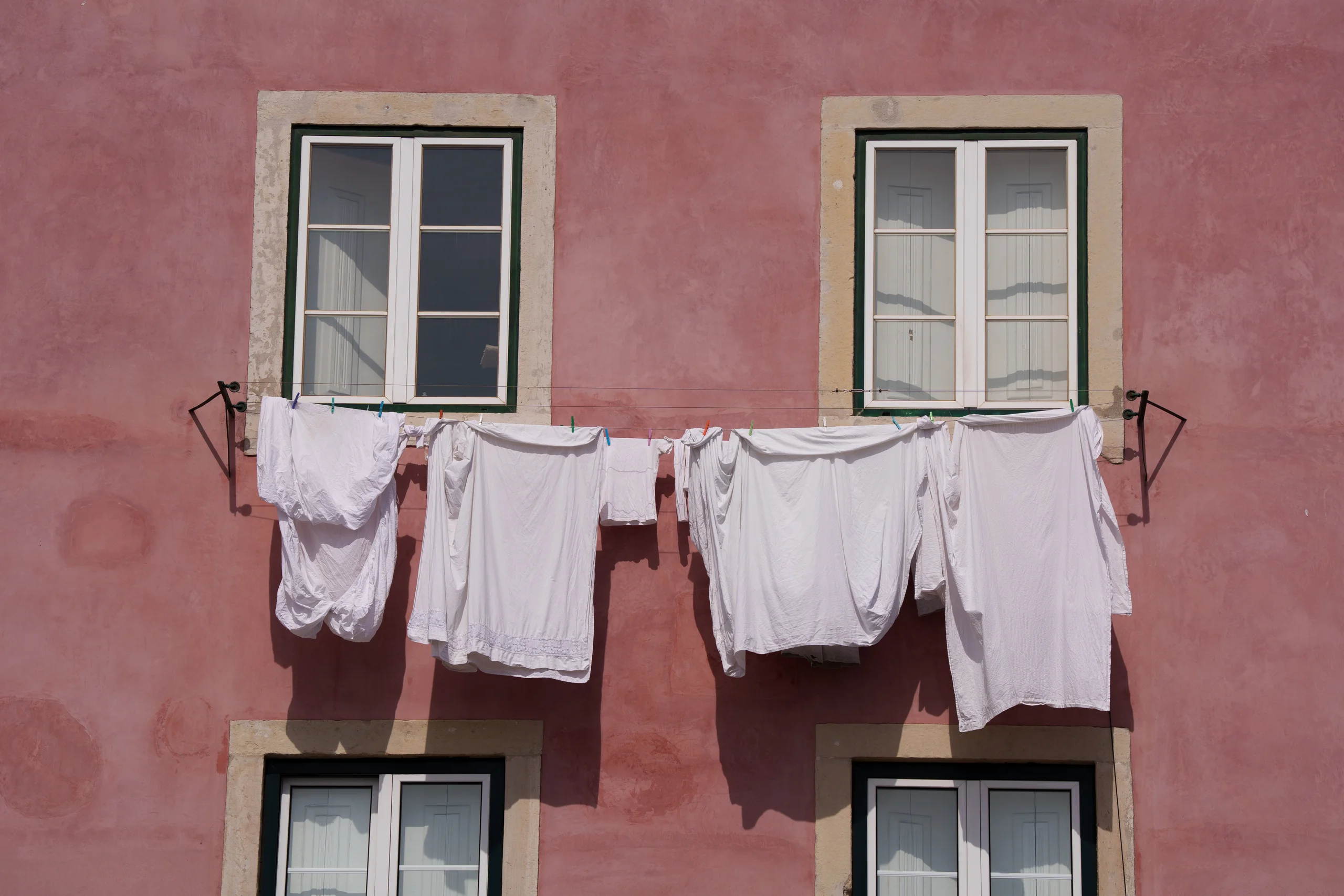 White laundry hangs on a line strung between two windows of a salmon-pink Lisbon facade.