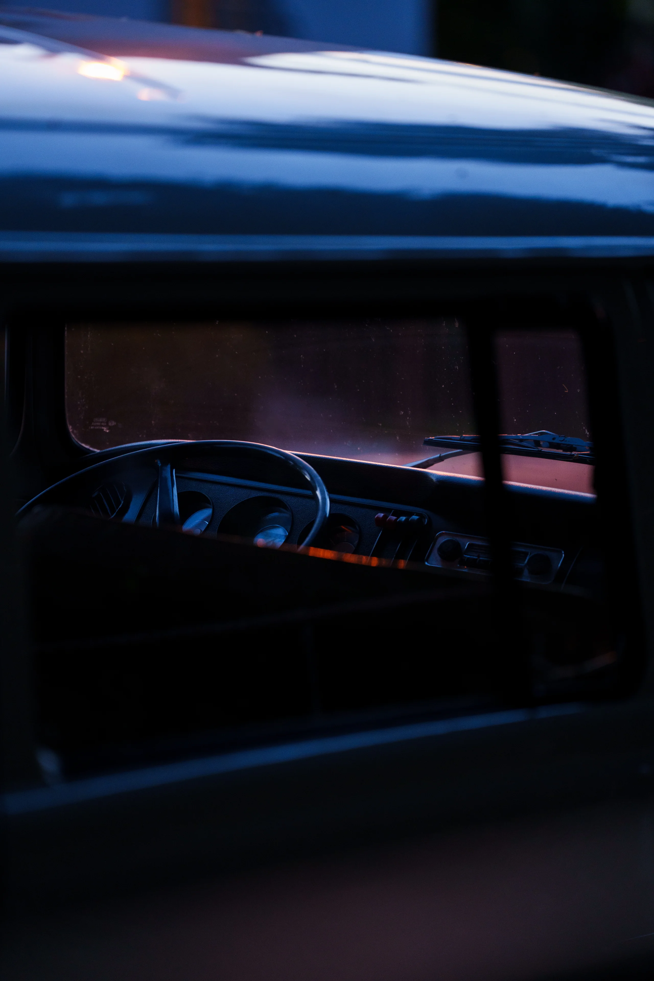 A vintage car interior is glimpsed through an open window at dusk, cool blue tones playing across the bodywork while the dashboard glows warmly within.