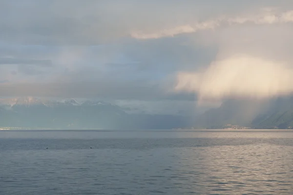 Rain veils the distant Alps across Lake Geneva, soft curtains of cloud trailing over the water under a hazy evening light.