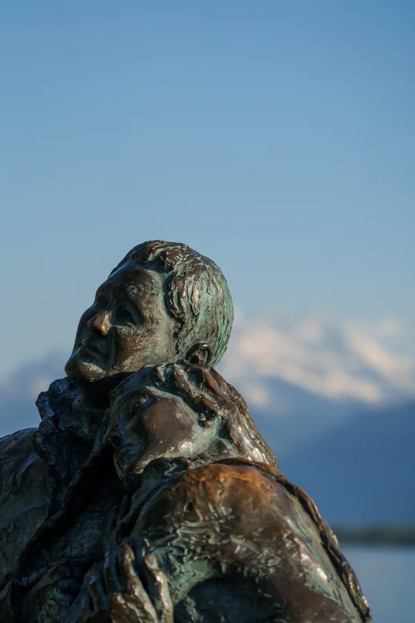 A weathered bronze statue of two embracing figures, its green-and-rust patina catching the light, set against the soft snow-capped Alps.