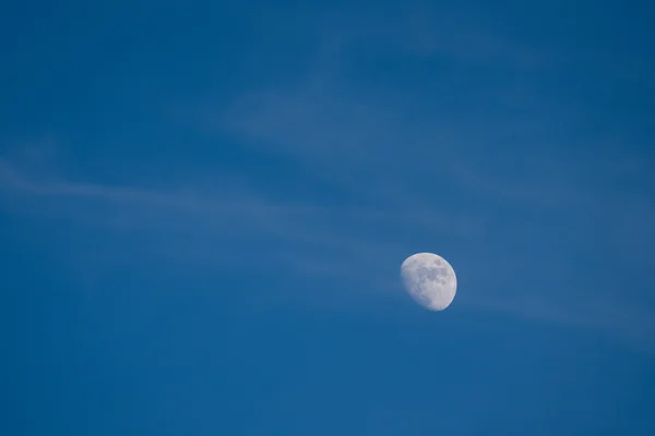 The waxing gibbous Moon hangs in a deep blue afternoon sky, thin cirrus streaks drifting around it.
