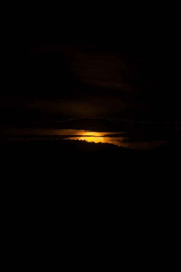 The setting sun glows orange as it slips behind a dark, forested ridge beneath a heavy band of cloud.