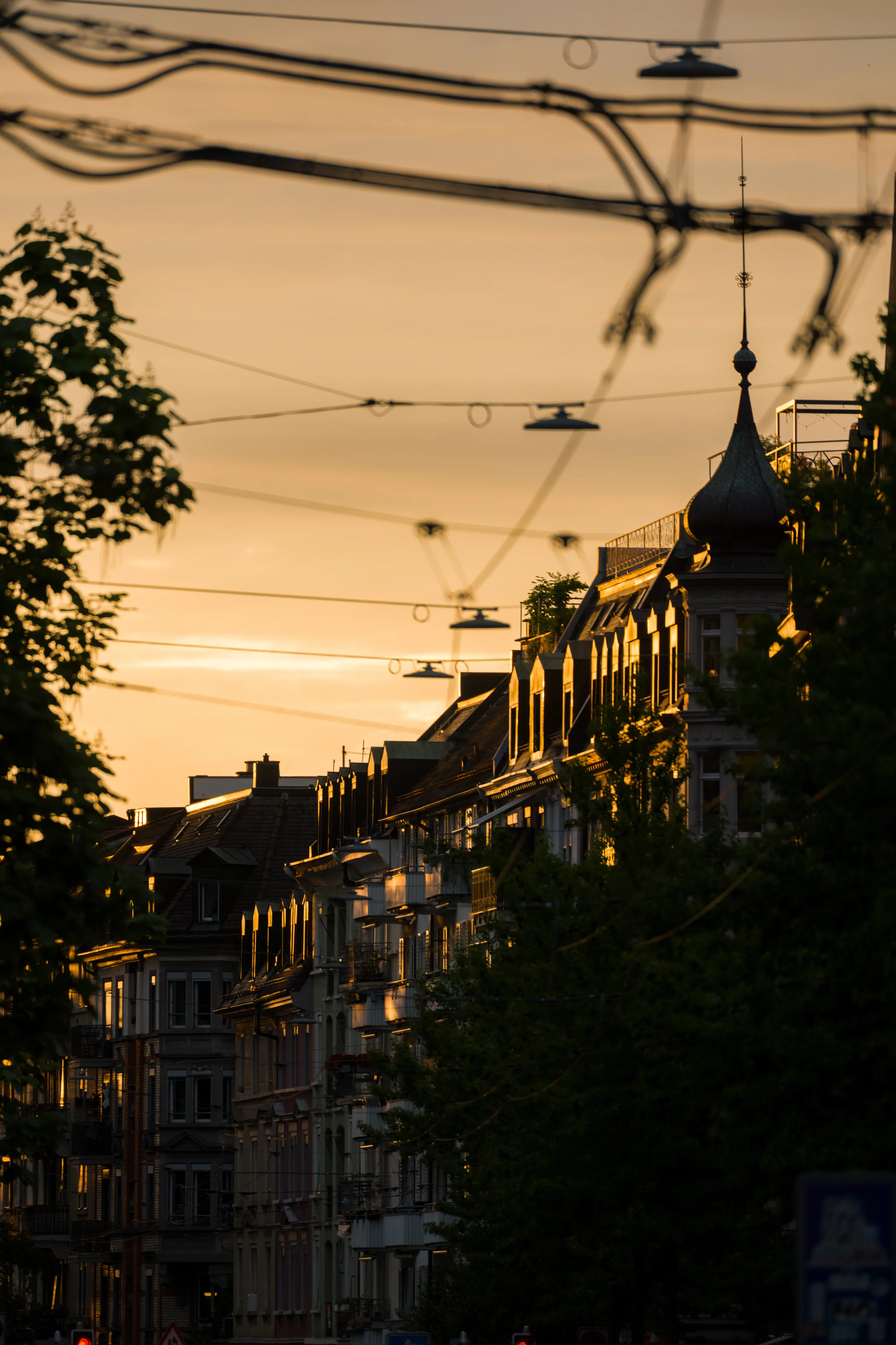 Golden evening light catches the facades of a row of buildings along a Zurich avenue, an ornate turret rising above the rooftops as tram wires crisscross the sky.