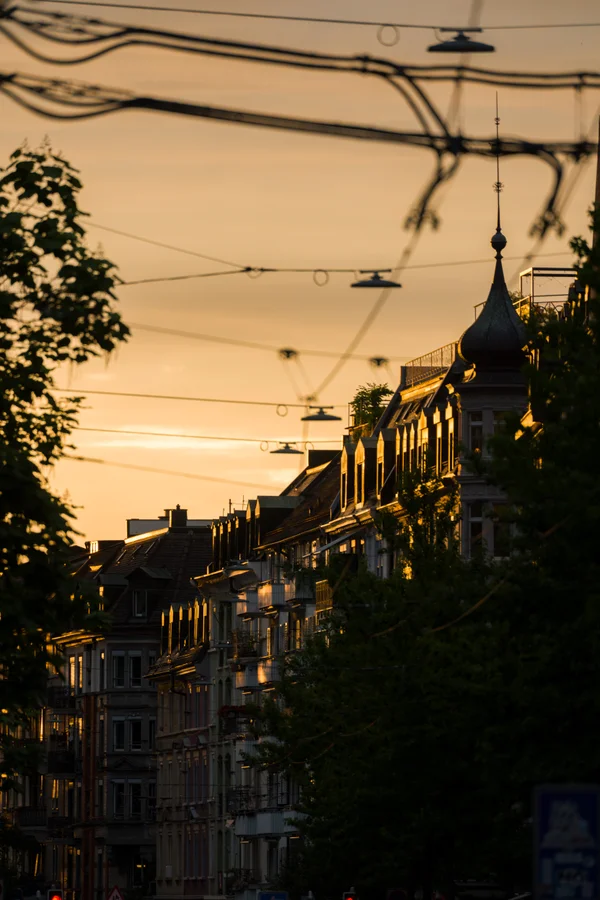 Golden evening light catches the facades of a row of buildings along a Zurich avenue, an ornate turret rising above the rooftops as tram wires crisscross the sky.