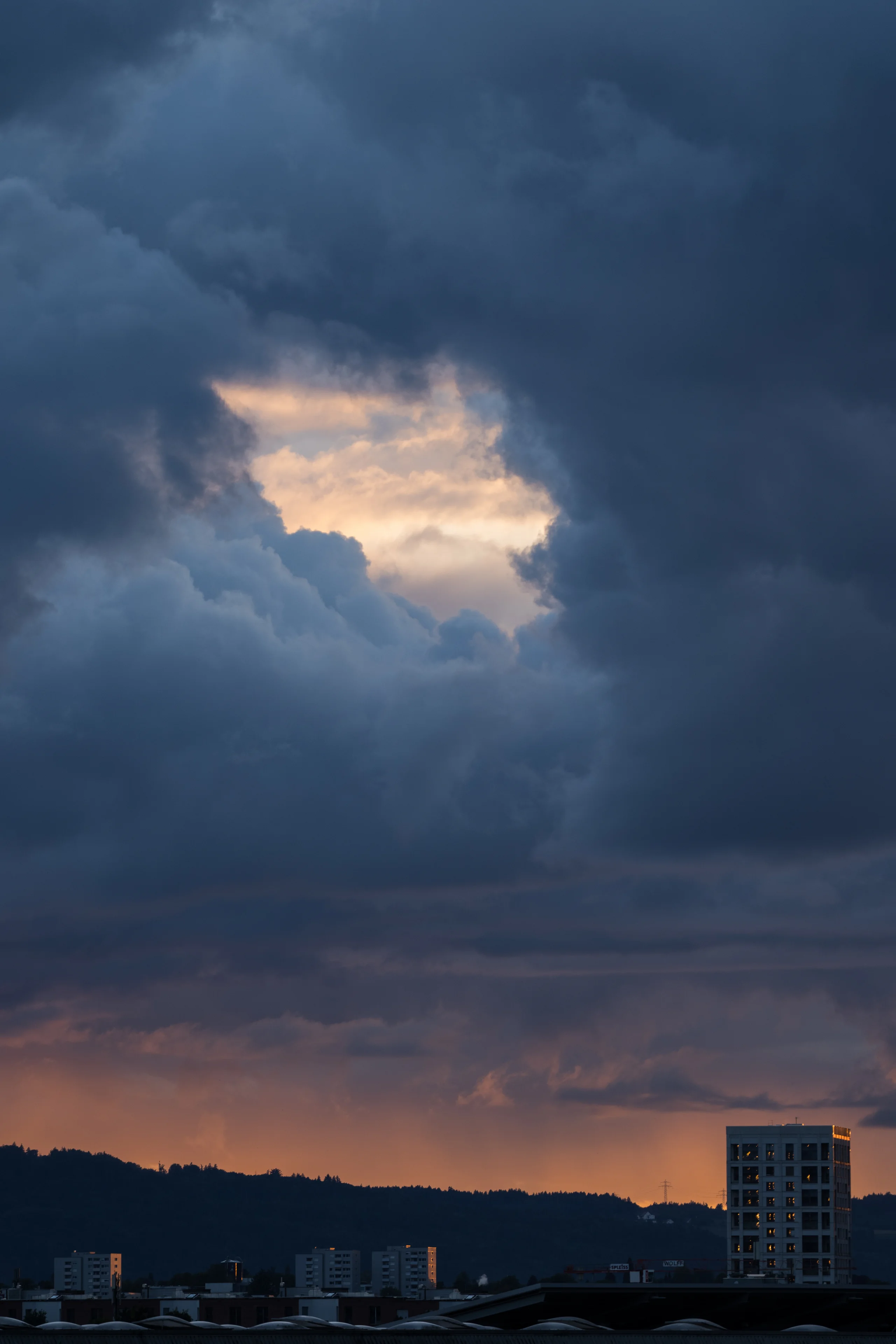 Dark storm clouds dominate the sky as a bright sunlit patch breaks through the center, with distant rain streaks falling over a city skyline silhouetted below.