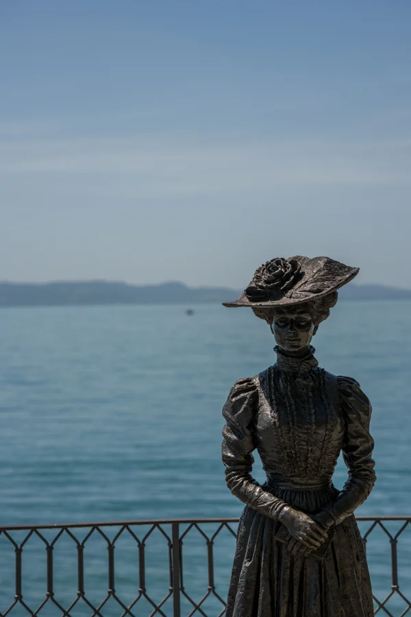 A bronze statue of a woman in a Victorian dress and wide rose-trimmed hat stands at a lakeside railing, the calm waters of the lake stretching to a distant shore behind her.