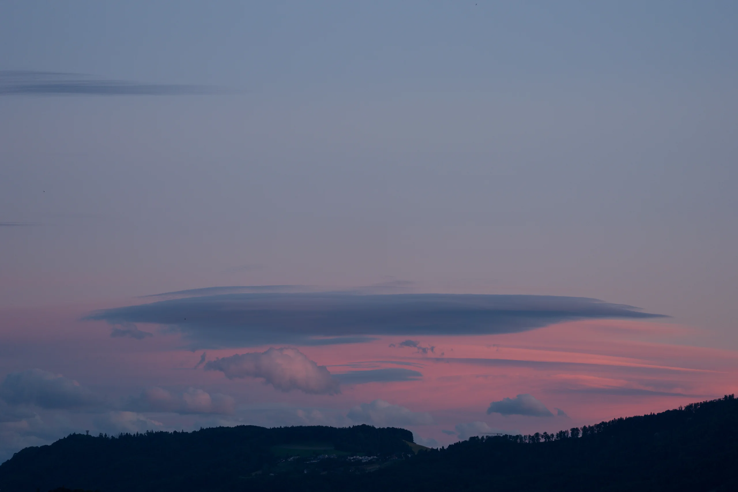 A flat, lens-shaped lenticular cloud hovers above a forested ridge, lit by warm pink and coral tones from the sunset sky.