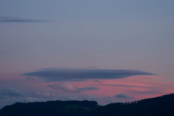 A flat, lens-shaped lenticular cloud hovers above a forested ridge, lit by warm pink and coral tones from the sunset sky.
