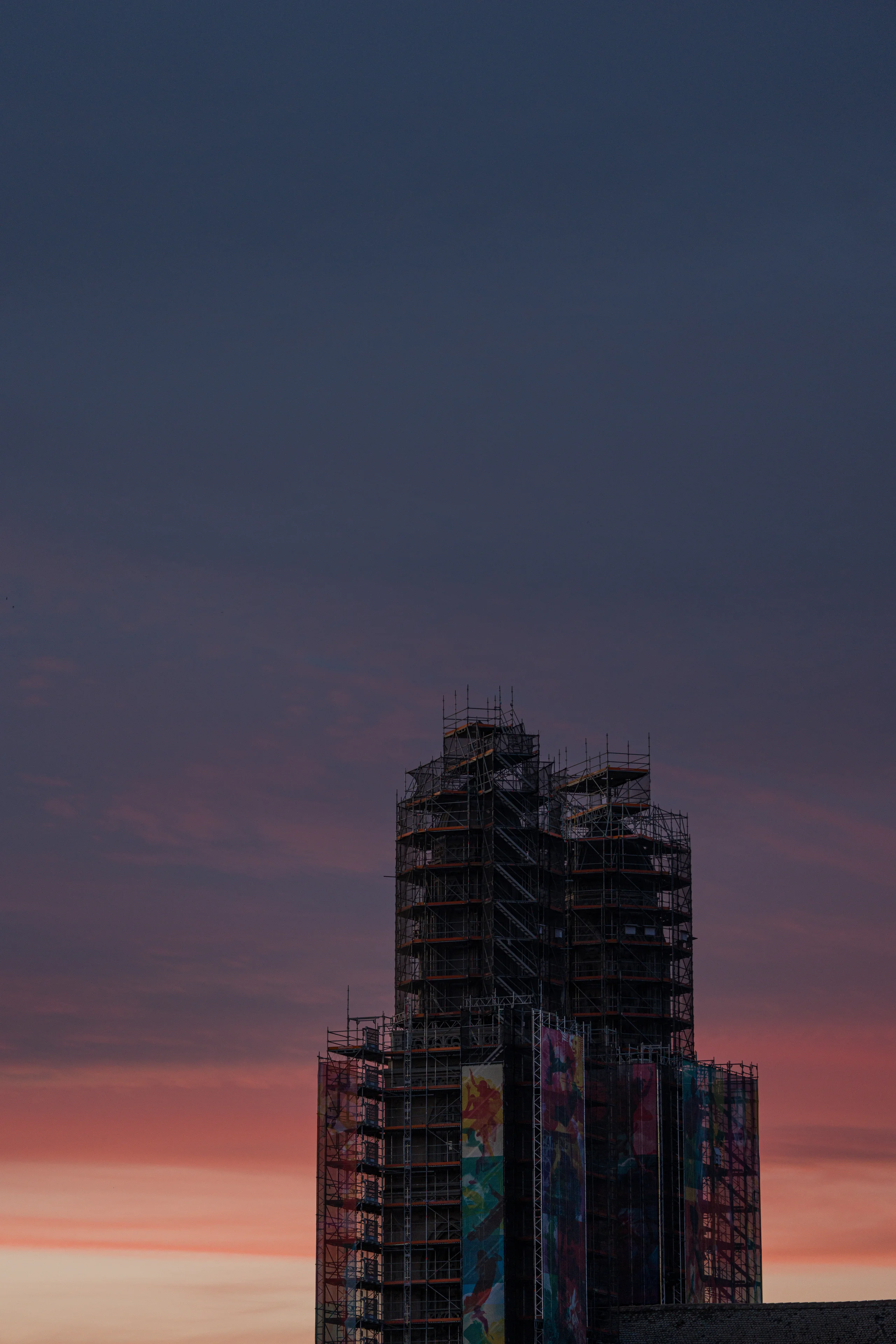 Grossmünster wrapped in scaffolding and colorful mural panels, seen from Quaibrücke against a soft gradient sunset sky fading from pink to deep blue.