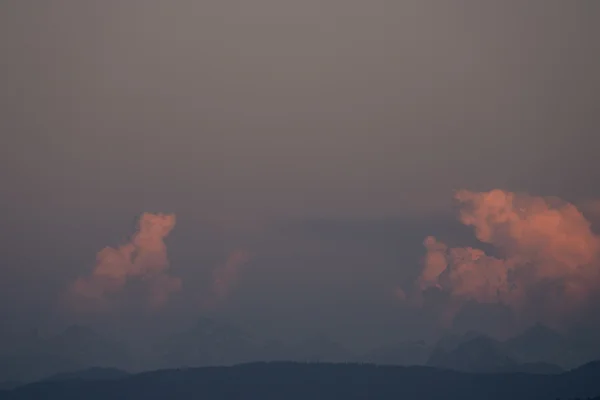 A glowing pink cumulus cloud catches warm light above hazy snow-capped Alps silhouetted along the bottom, with a soft mauve sky stretching above.