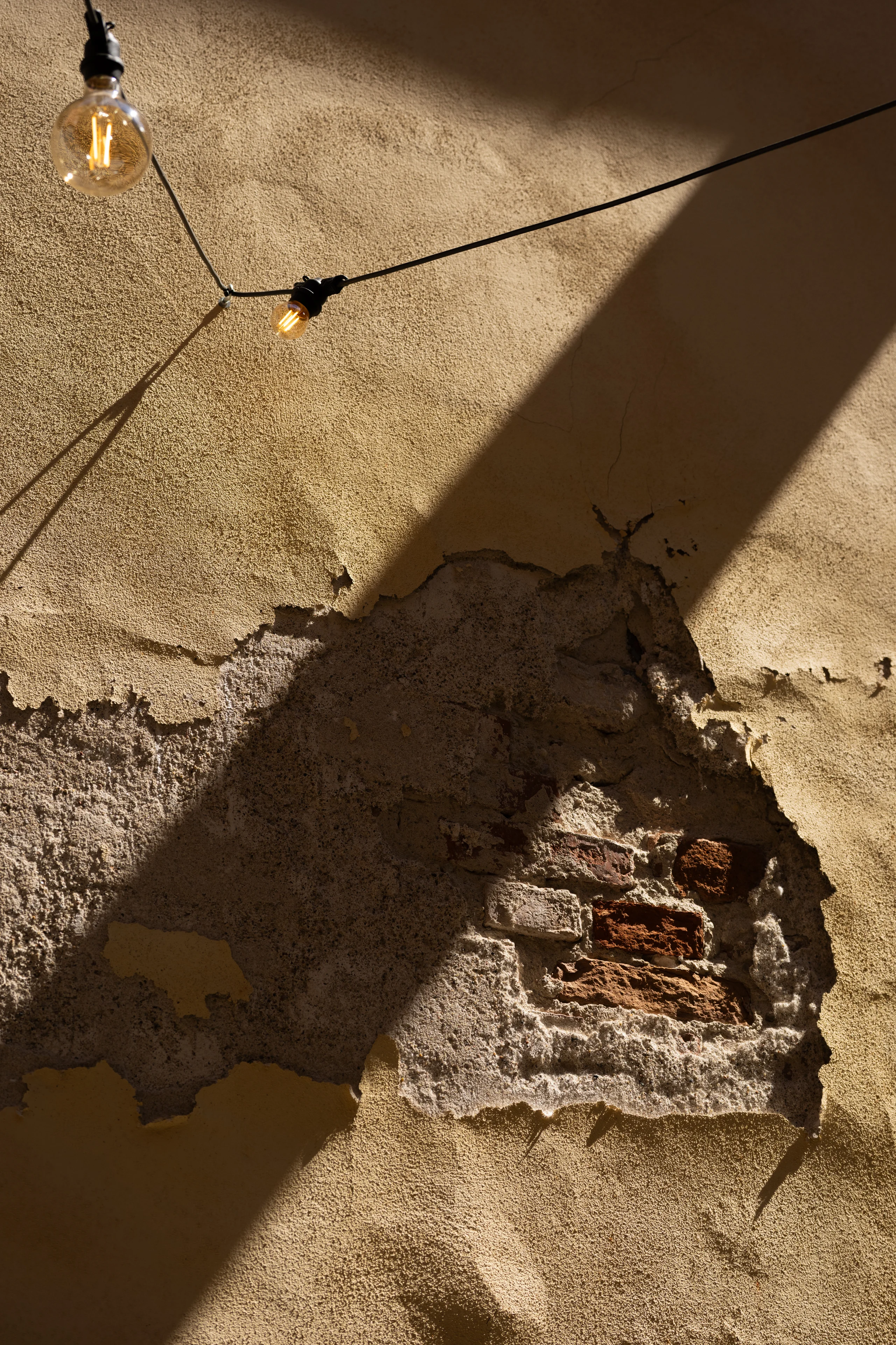 Diagonal sunlight cuts across a textured stucco wall where a patch of crumbling plaster reveals worn brick beneath, with two vintage Edison-style bulbs hanging on a wire above.