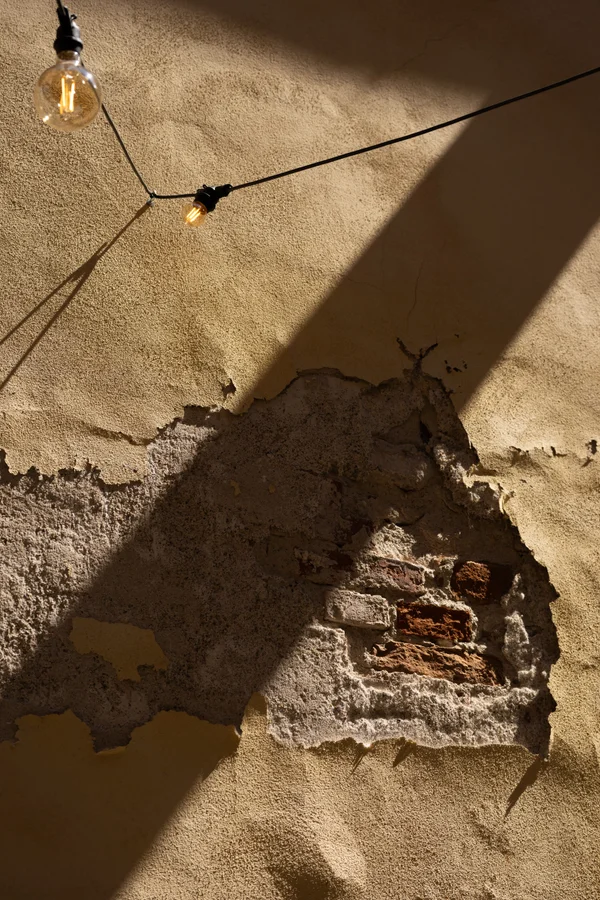 Diagonal sunlight cuts across a textured stucco wall where a patch of crumbling plaster reveals worn brick beneath, with two vintage Edison-style bulbs hanging on a wire above.