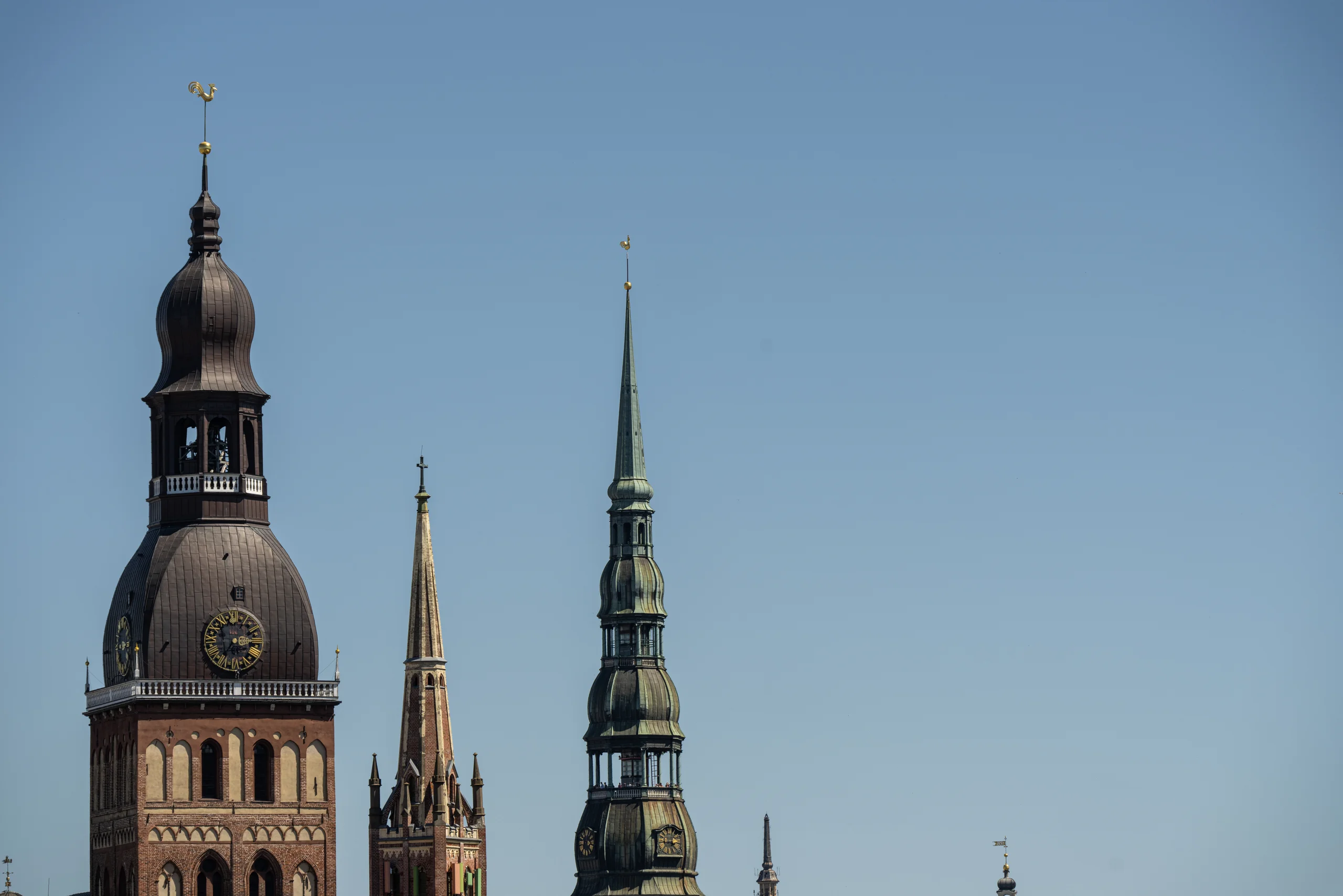 Historic Rīga Old Town spires rise against a clear blue sky, with the bulbous cupola of Rīgas Doms on the left, a smaller pointed brick spire between them, and the tall green spire of Sv. Pētera baznīca on the right.