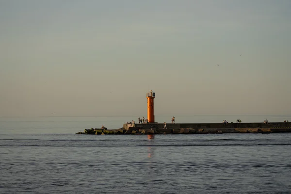 A red lighthouse stands on a long rocky breakwater extending into a calm sea, with small figures walking along it under warm evening light and seabirds drifting in the distance.