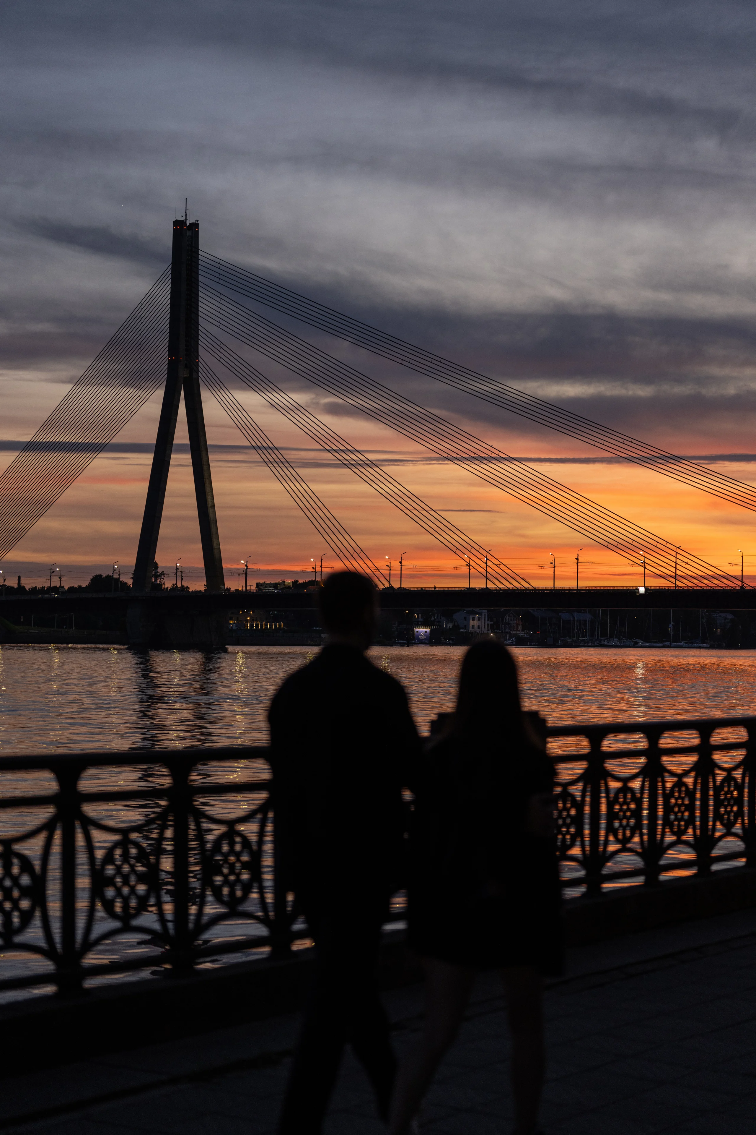 A couple stands silhouetted at a decorative railing watching the Vanšu tilts cable-stayed bridge across the Daugava, with warm sunset tones reflected on the river.