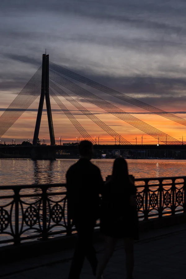 A couple stands silhouetted at a decorative railing watching the Vanšu tilts cable-stayed bridge across the Daugava, with warm sunset tones reflected on the river.