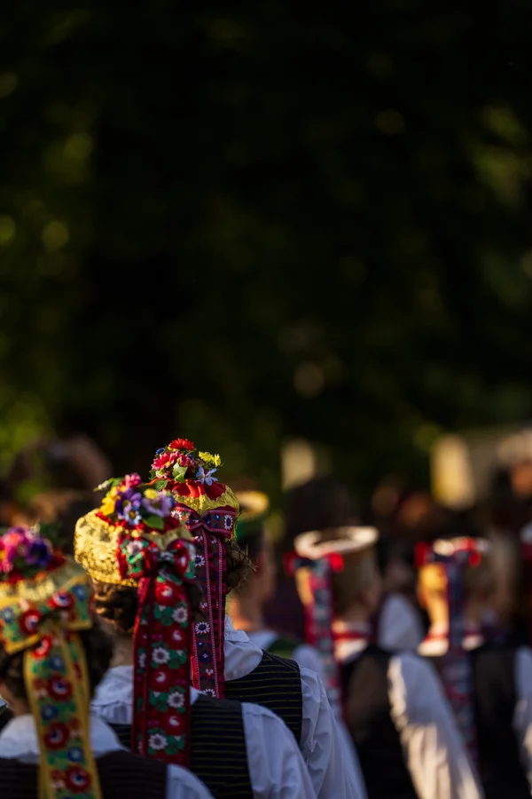 People in traditional Latvian folk dress, seen from behind, with vibrant floral crowns and long embroidered ribbons trailing down their backs in deep reds, yellows, and pinks.