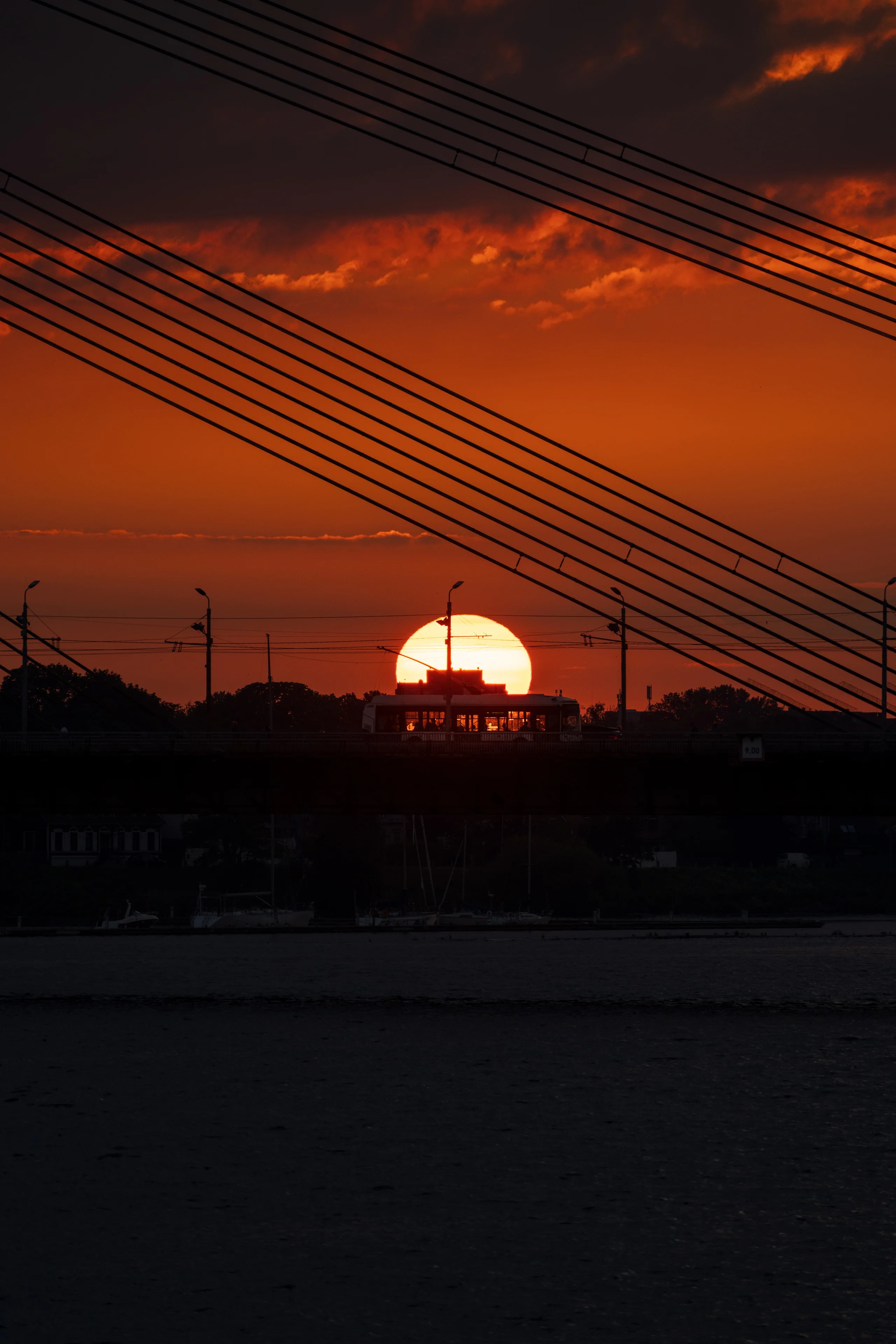 The setting sun glows massive and orange, perfectly framed behind a bus crossing the bridge, with diagonal cables of the Vanšu tilts cutting across the upper frame against a deep red sky.