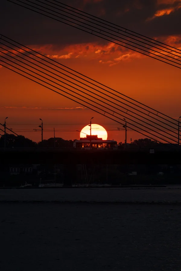The setting sun glows massive and orange, perfectly framed behind a bus crossing the bridge, with diagonal cables of the Vanšu tilts cutting across the upper frame against a deep red sky.