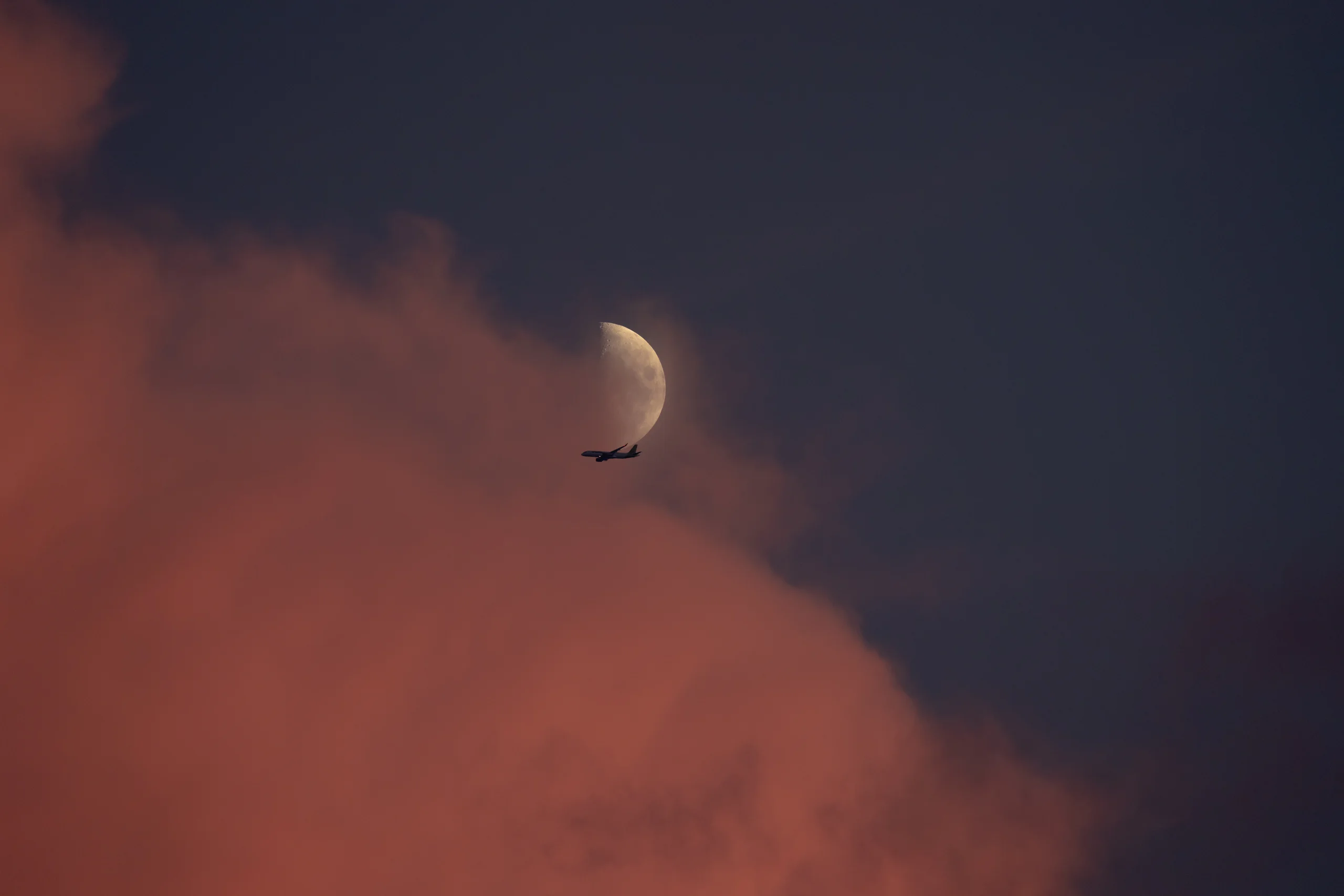A waxing half moon hangs in the upper sky as a passenger airplane silhouetted glides just below it, with bright coral-pink afterglow clouds catching the last light below.
