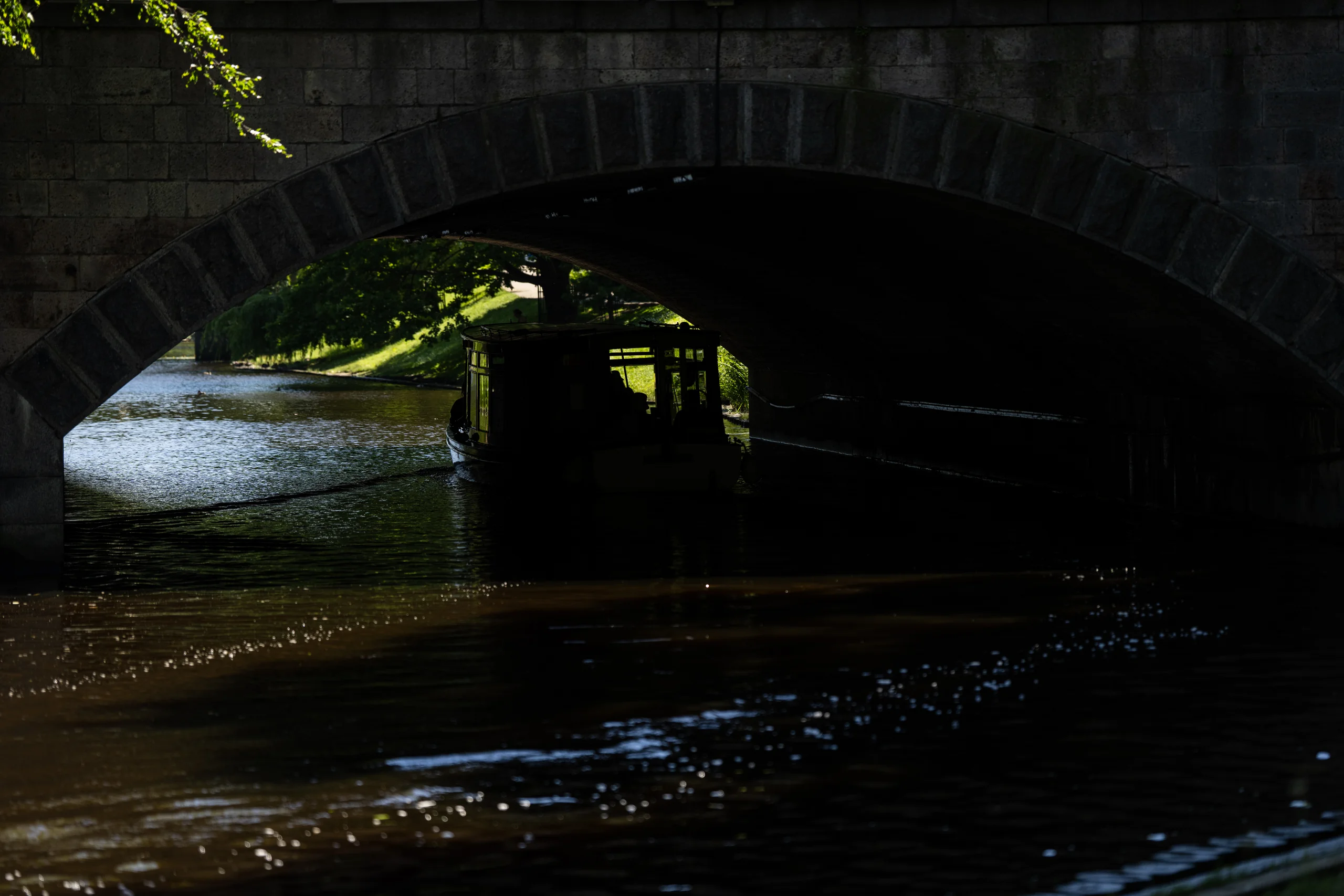 A covered boat glides beneath a stone arch bridge toward a sunlit grassy bank while light scatters across the dark water.