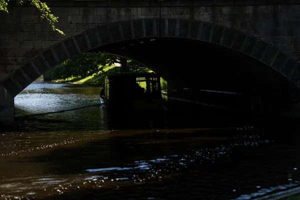 A covered boat glides beneath a stone arch bridge toward a sunlit grassy bank while light scatters across the dark water.