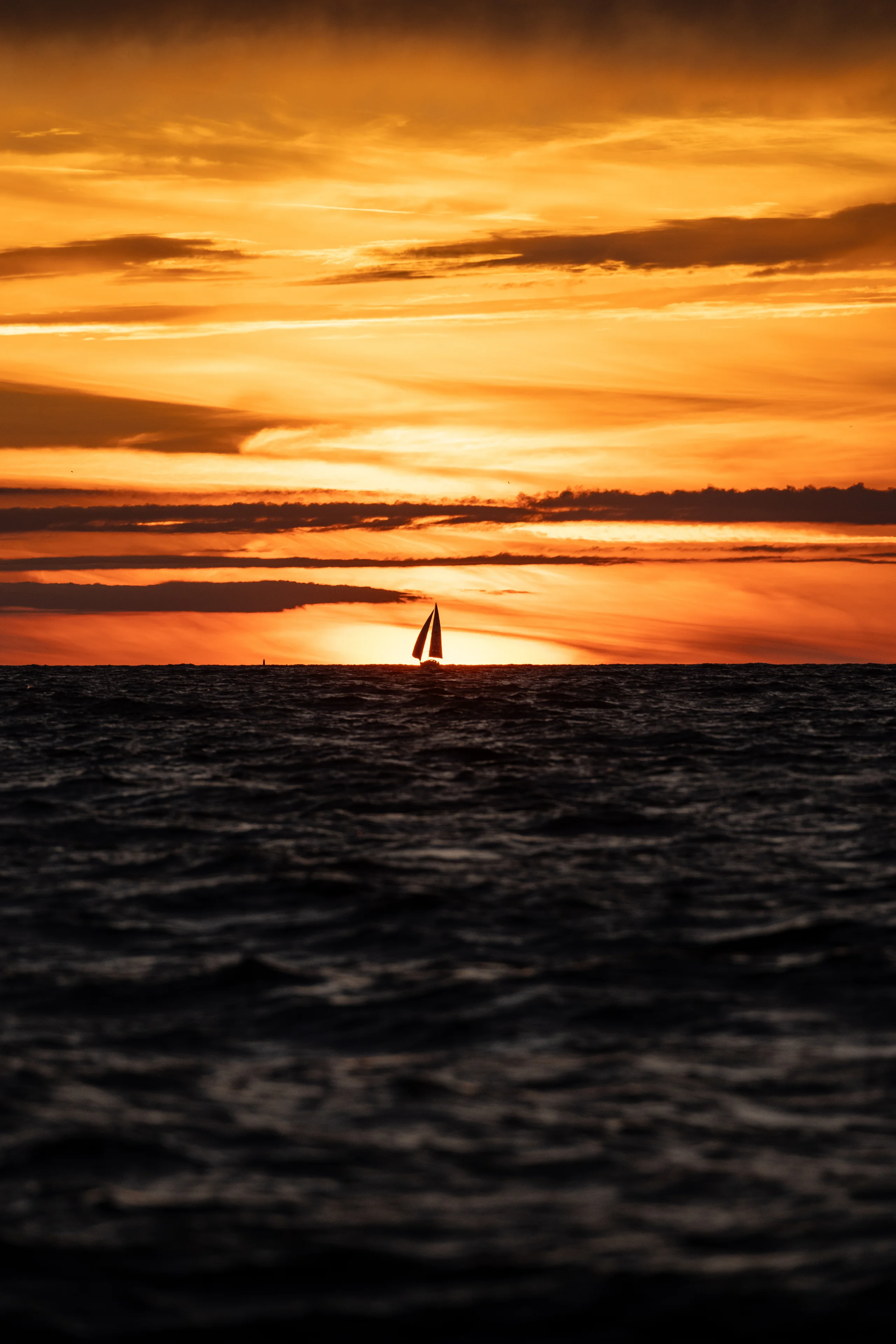 A sailboat rests on the horizon beneath a banded orange sky as dark water ripples across the foreground.