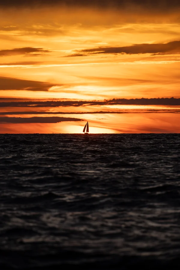 A sailboat rests on the horizon beneath a banded orange sky as dark water ripples across the foreground.