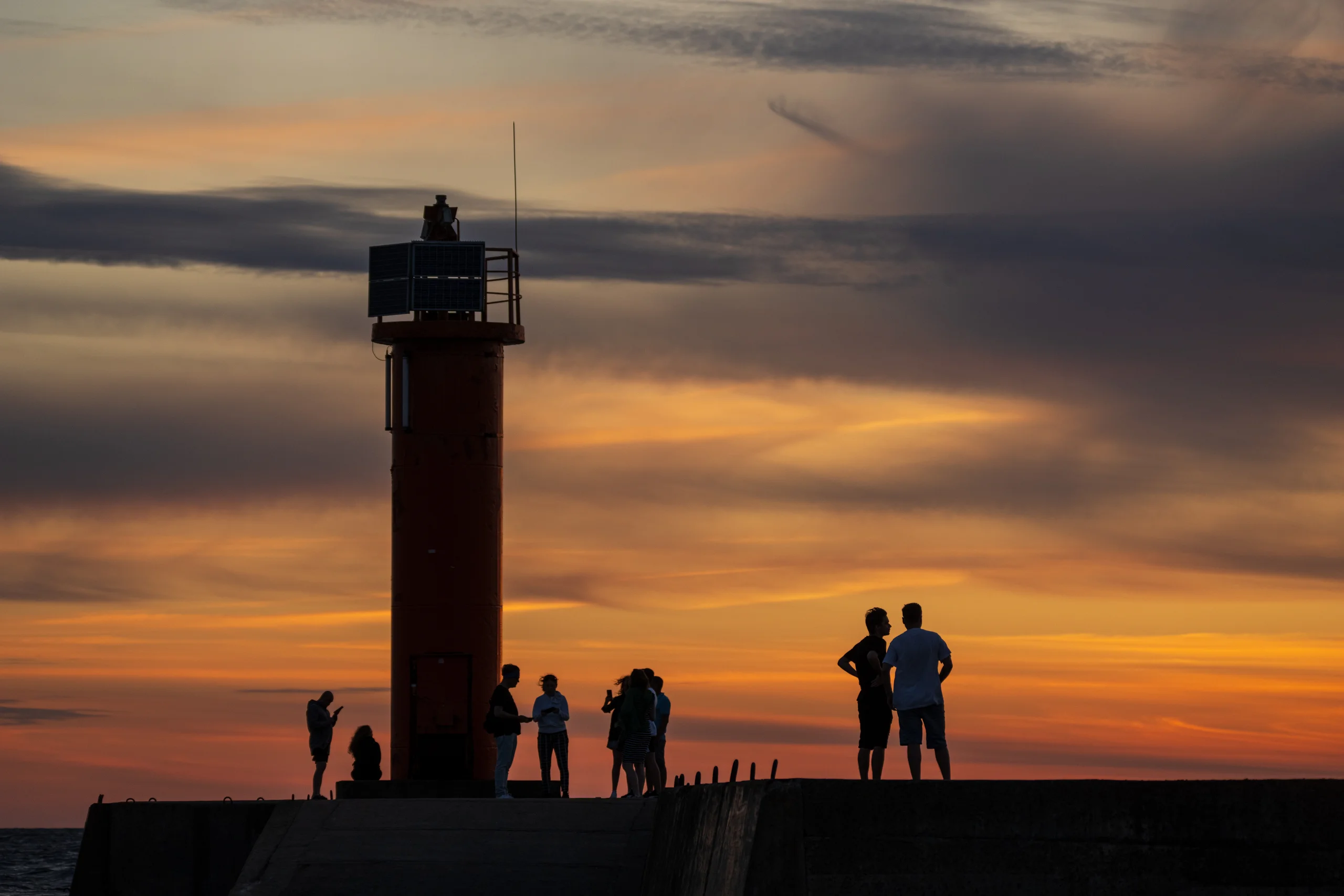 People stand along a stone breakwater beside a red lighthouse as an orange band of light lingers low beneath grey clouds.