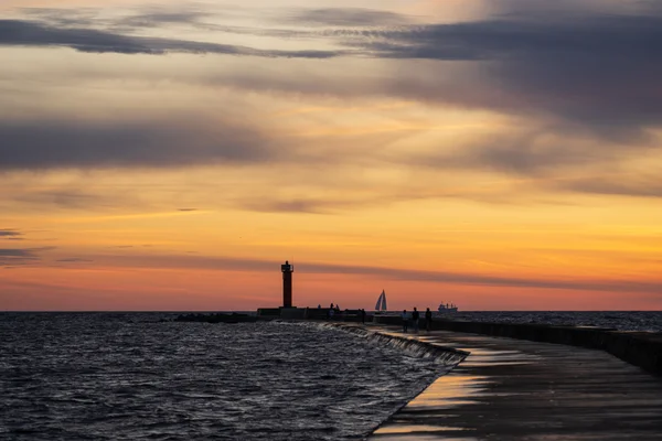 A curving stone breakwater leads toward a lighthouse as small figures stand along the pier beneath a fading orange sky.