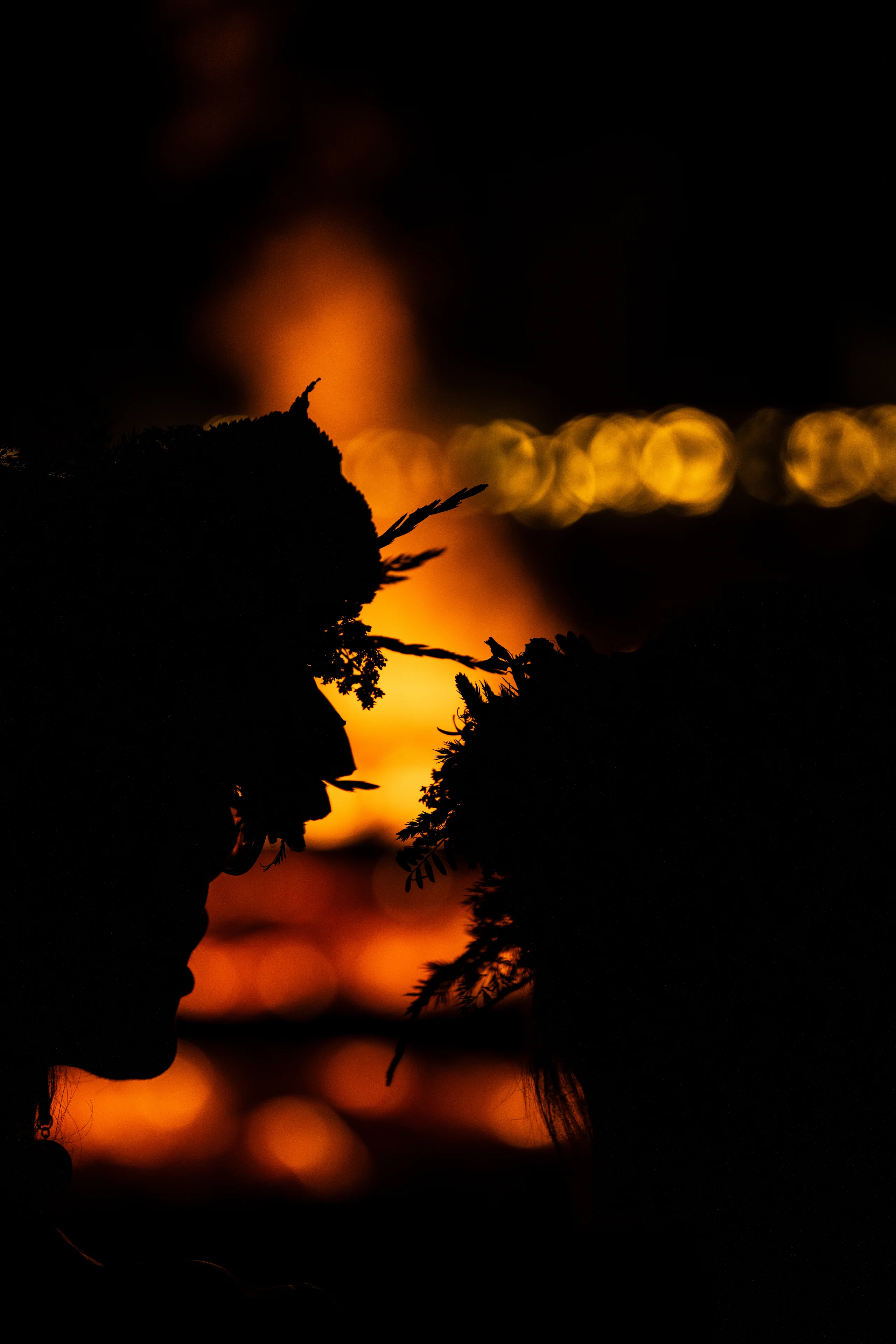Two people in floral crowns stand in profile, silhouetted against the orange glow of a Jāņi bonfire and rows of golden light.
