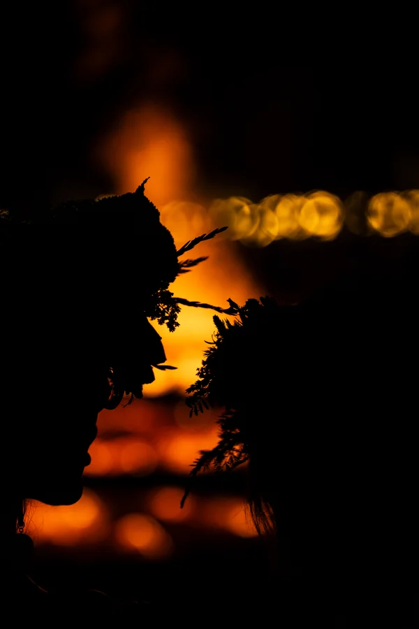 Two people in floral crowns stand in profile, silhouetted against the orange glow of a Jāņi bonfire and rows of golden light.