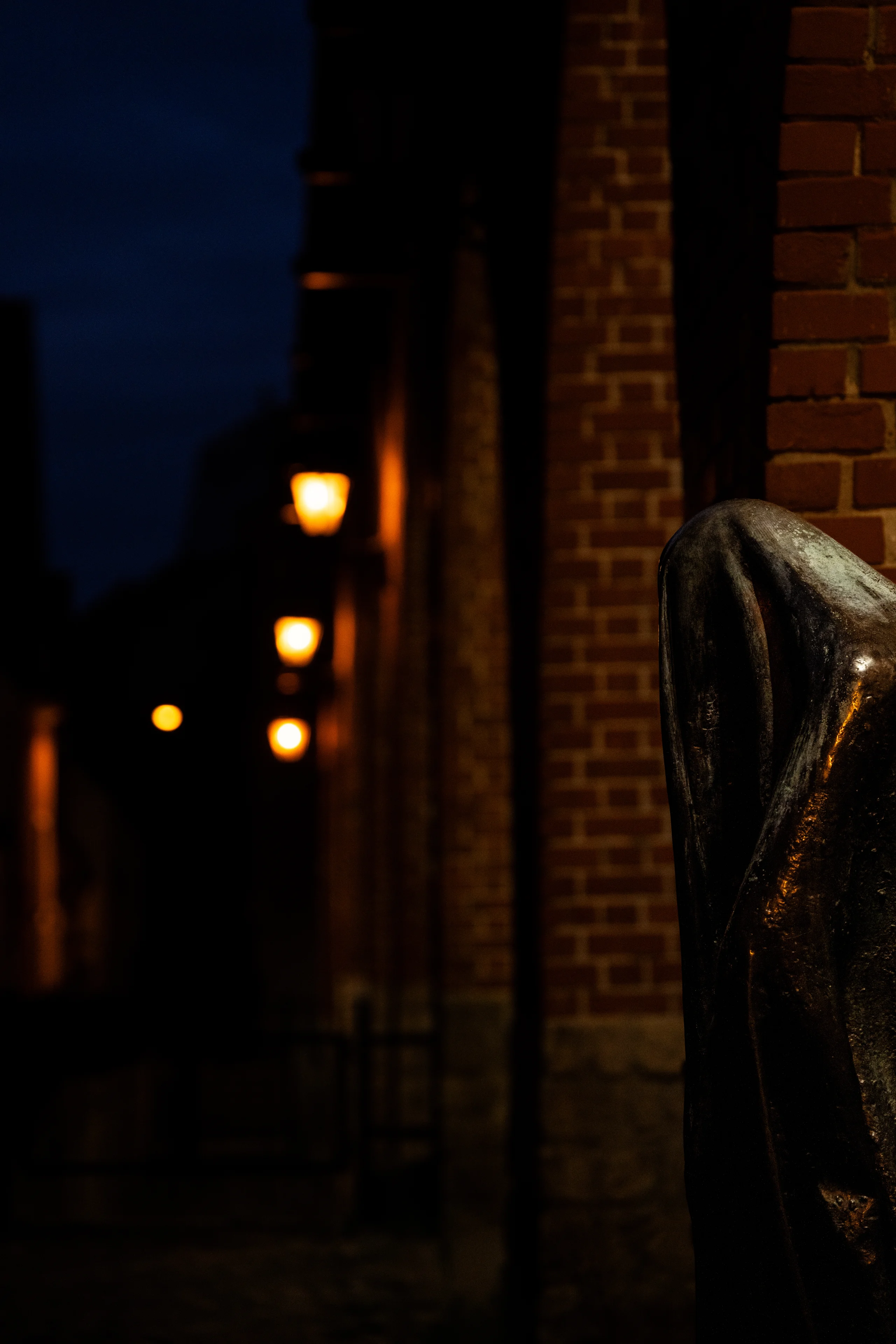 A cloaked bronze figure stands in shadow beside a brick wall as warm lanterns glow down a dark alley at night.