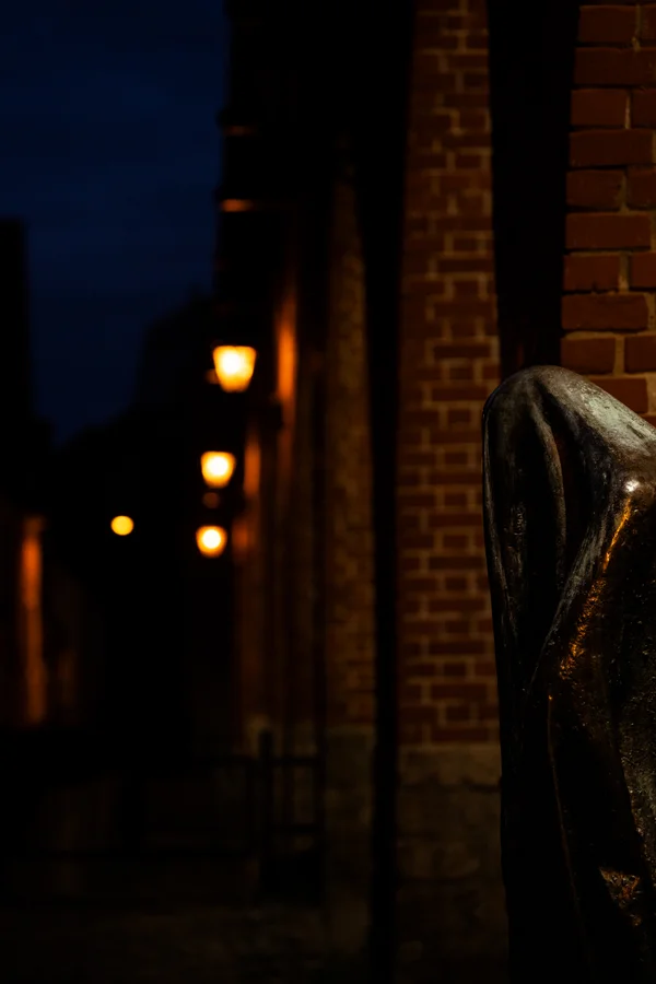 A cloaked bronze figure stands in shadow beside a brick wall as warm lanterns glow down a dark alley at night.