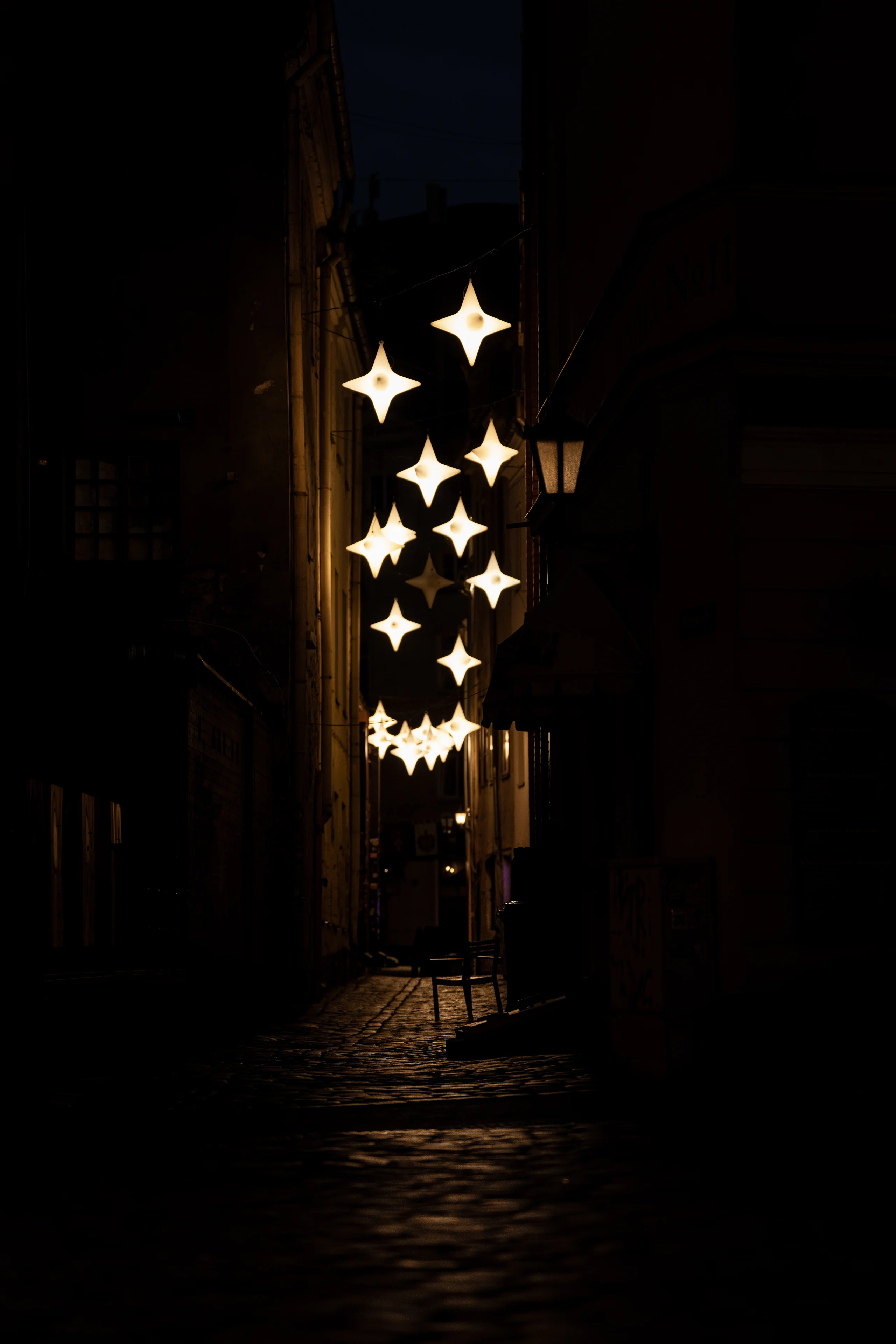 A vertical cluster of glowing star-shaped lanterns lights a narrow cobblestone alley at night above an empty chair on the stones.