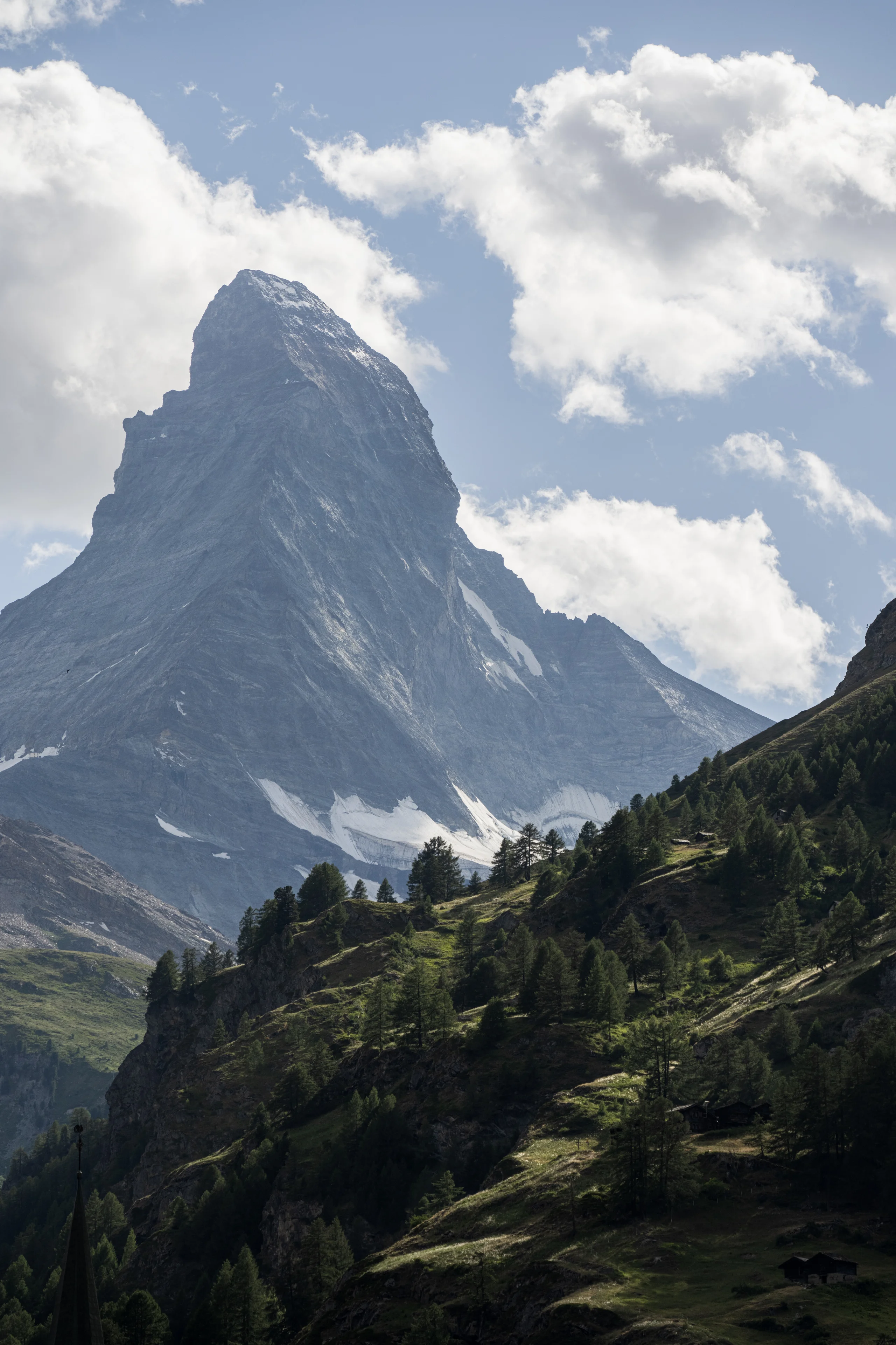The Matterhorn rises above forested slopes and scattered chalets under a blue sky with drifting clouds.