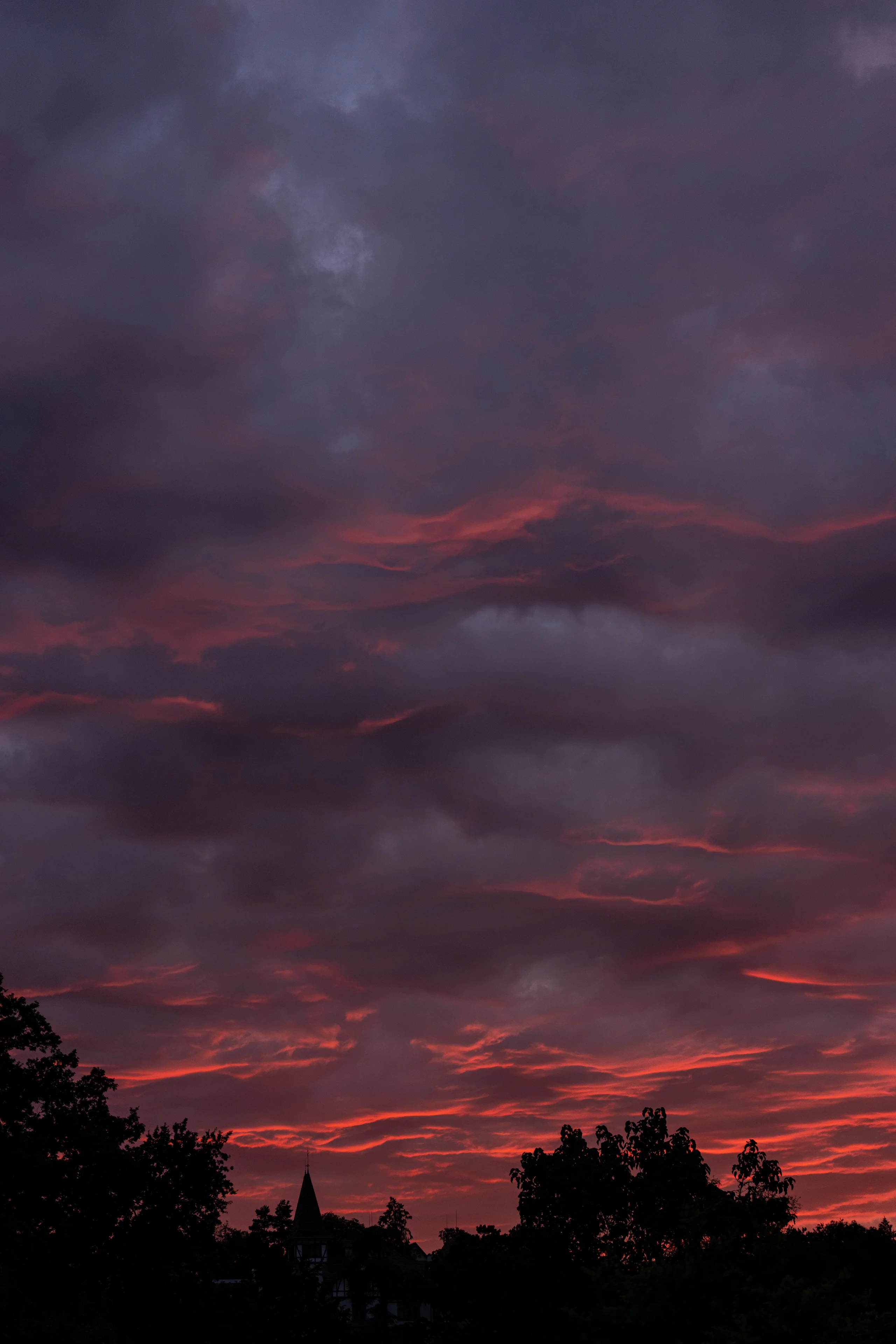 Deep purple clouds glow with red and coral afterglow above a silhouetted tree line and a small turreted building.