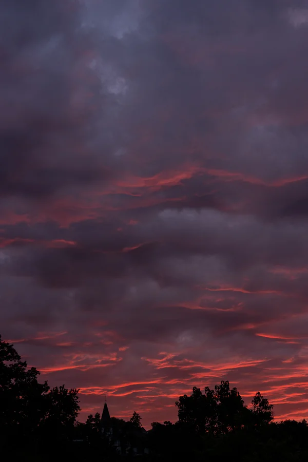 Deep purple clouds glow with red and coral afterglow above a silhouetted tree line and a small turreted building.